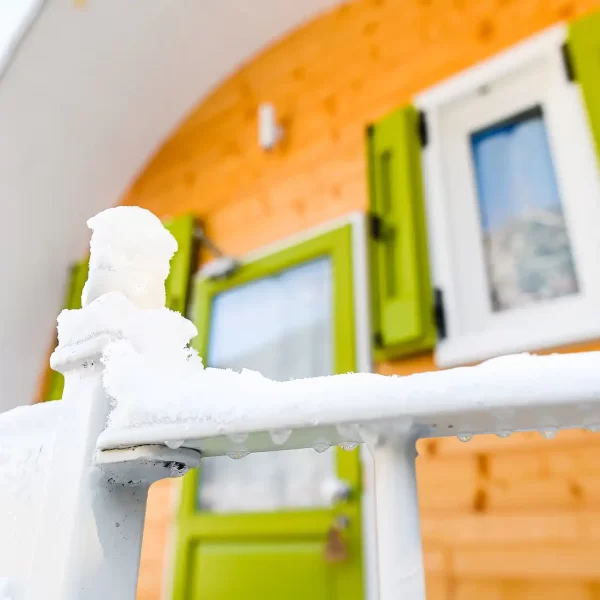 Verschneites Geländer vor einem kleinen Holzhäuschen mit grünen Fensterläden.